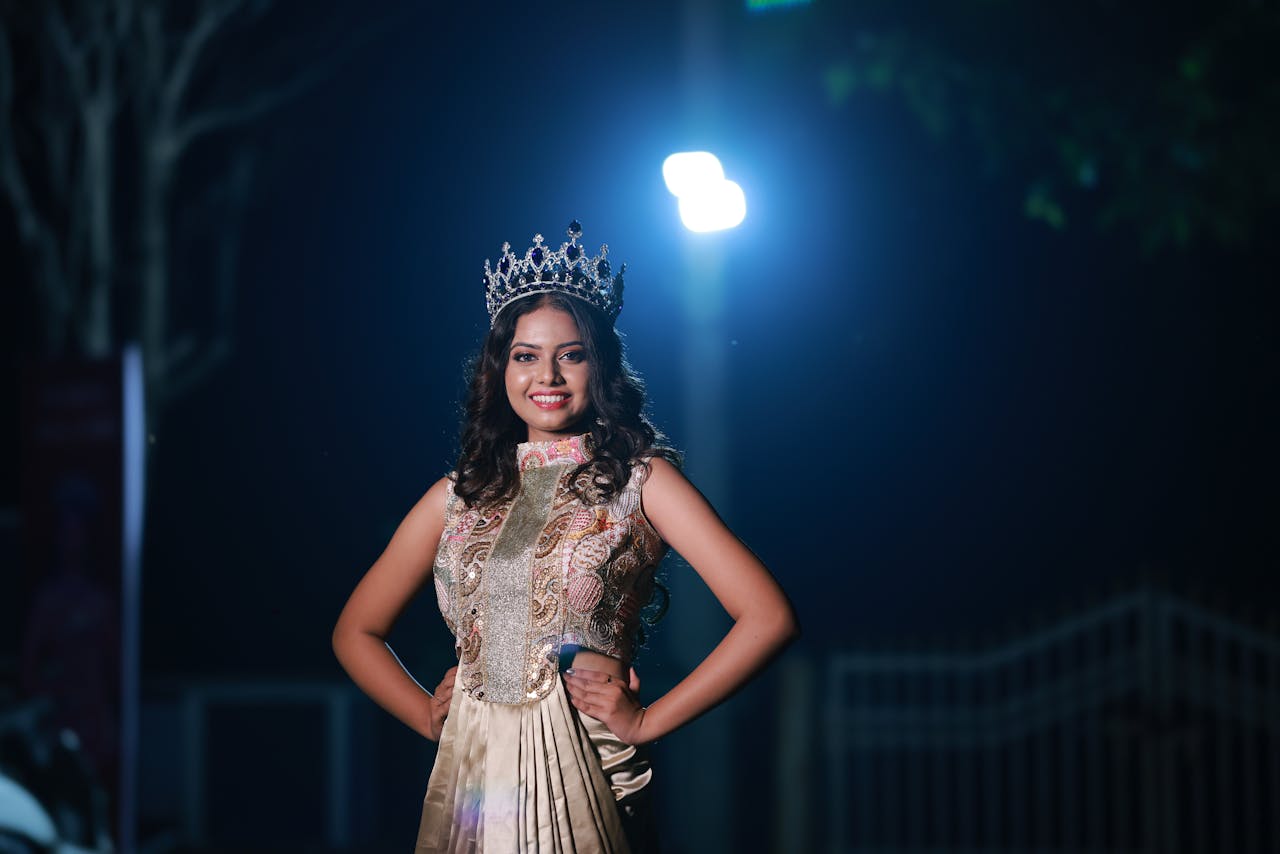 Beautiful woman wearing a crown and gown, posing confidently at night under dramatic lighting.